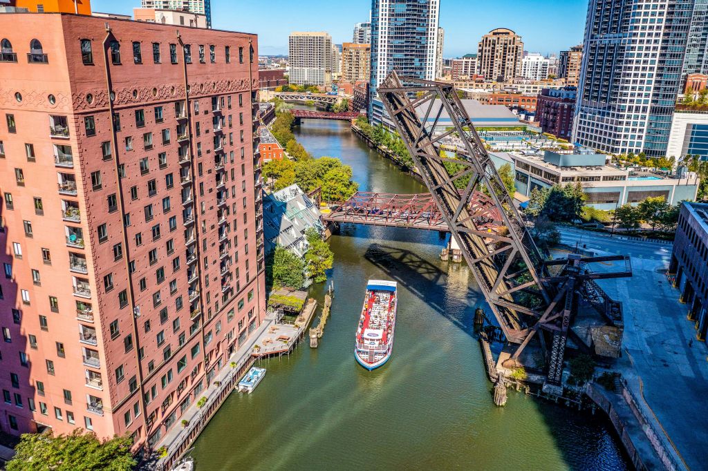 투어 보트가 있는 강, 양쪽에 있는 건물, 높이 솟은 녹슨 다리, 고층 빌딩을 배경으로 한 공중 전망.