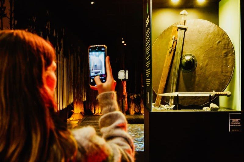 한 사람이 조명이 어두운 박물관에 전시된 칼과 방패가 있는 역사적인 전시물의 사진을 찍고 있습니다.