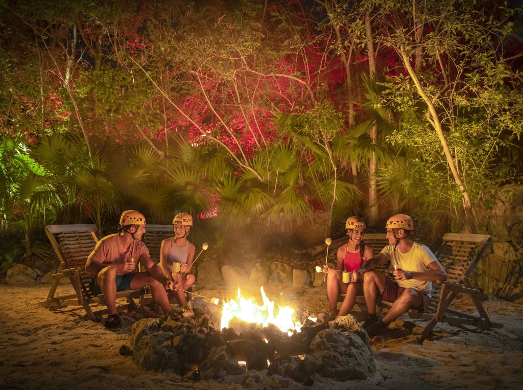 헬멧을 쓴 네 명이 야간에 형형색색의 조명이 켜진 나무에 둘러싸인 모닥불에 구운 마시멜로 옆에 앉아 있습니다.