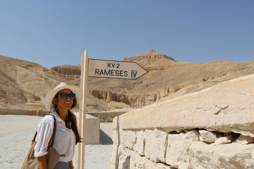 선글라스와 모자를 쓴 여성이 언덕이 있는 사막 풍경에서 "KV.2 Ramesses IV" 방향 표지판 옆에서 미소를 짓고 있습니다.