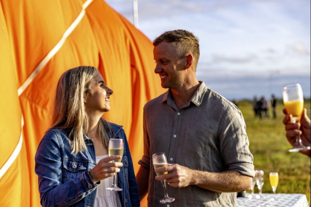 한 남녀가 와인 잔을 들고 서로에게 미소를 짓고 있습니다. 배경에는 커다란 주황색 구조물이 있습니다.