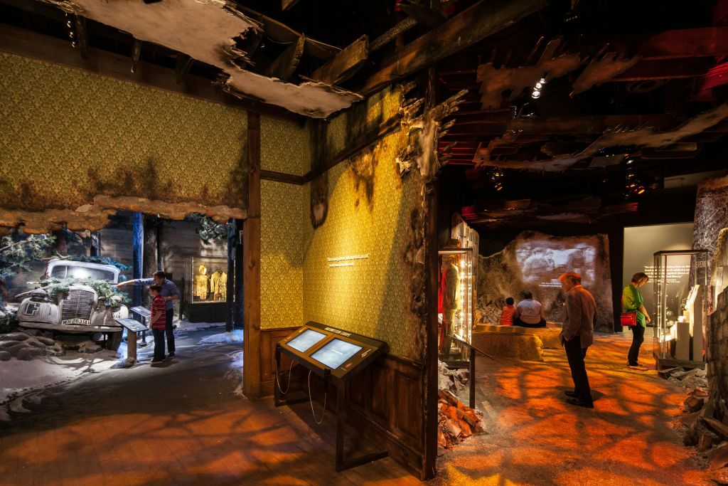 새까맣게 그을린 벽과 손상된 천장, 인터랙티브 스크린, 디스플레이를 보는 사람들의 모습을 보여주는 박물관 전시 장면.