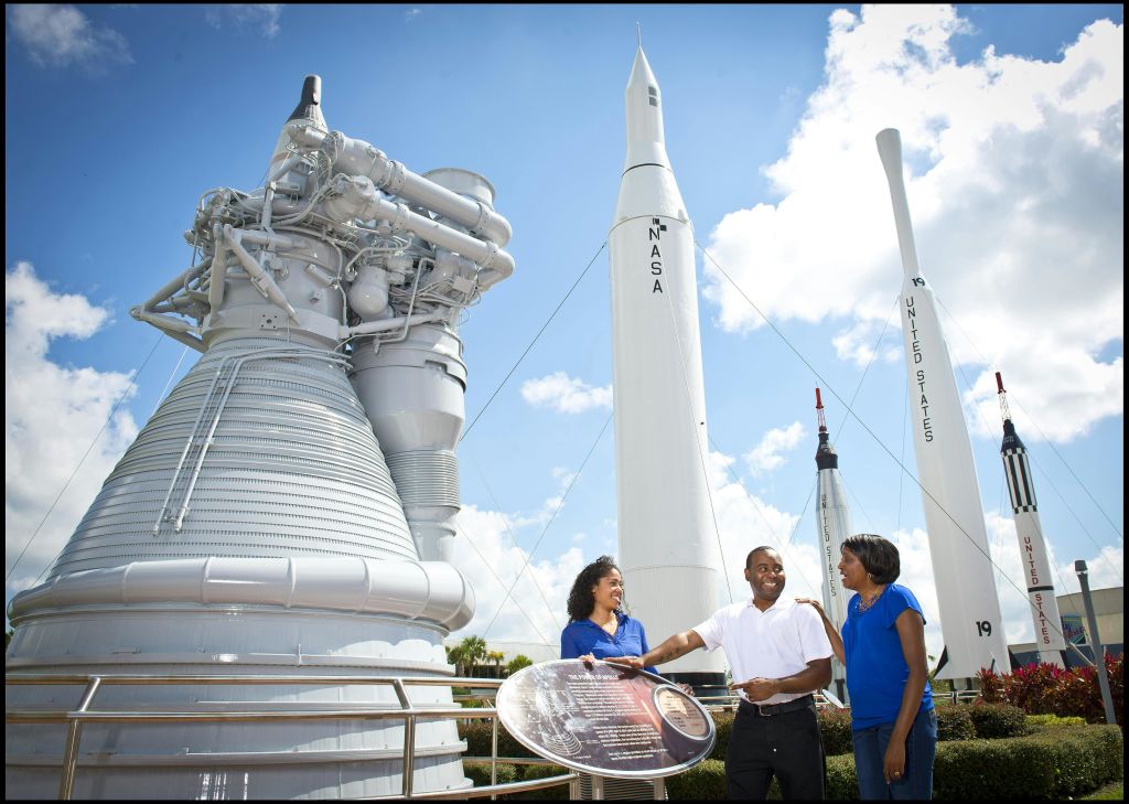 부분적으로 흐린 하늘 아래 NASA 시설에서 세 사람이 로켓과 엔진 디스플레이 근처에서 웃으며 서 있습니다.
