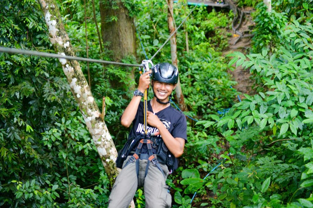 헬멧과 하네스를 착용한 채 울창하고 푸른 숲을 가로질러 집라인을 그리며 웃고 있는 사람.