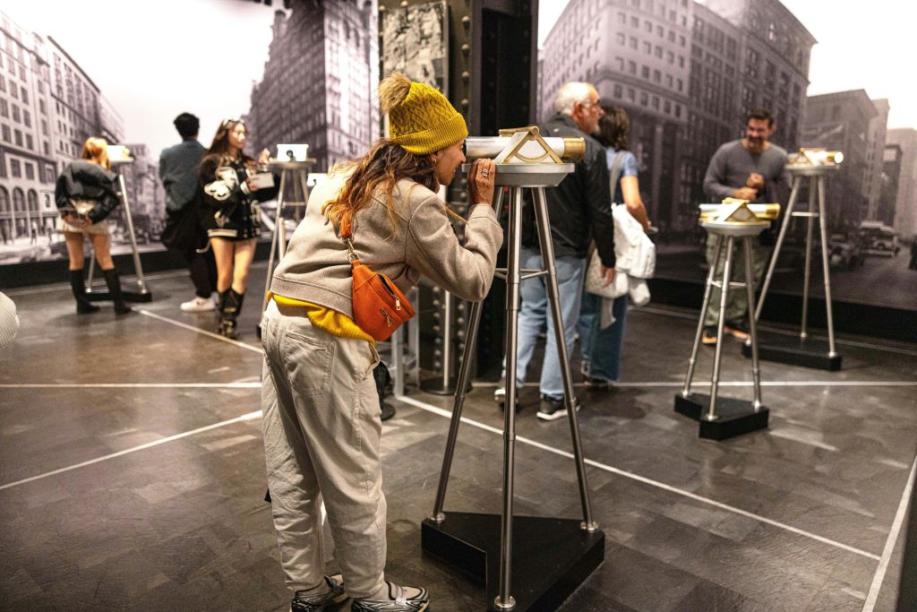 도시 경관을 배경으로 한 박물관 같은 공간에서 쌍안경 모양의 장치를 갖춘 전시물과 교류하는 사람들.