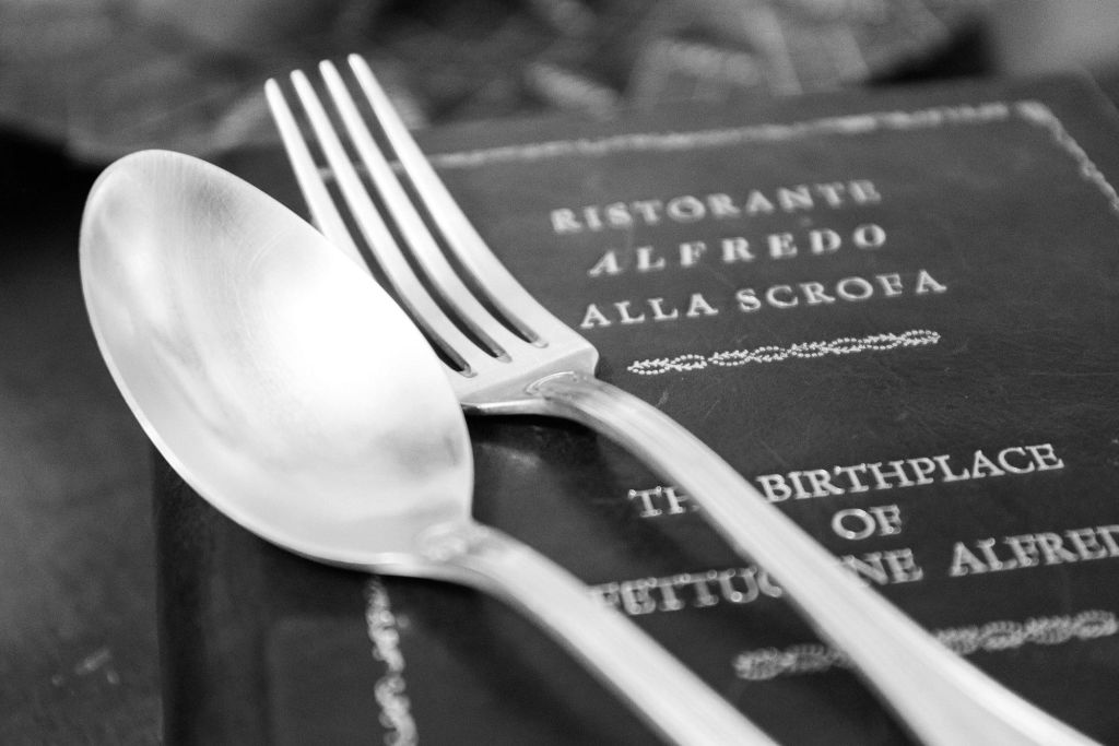 "RISTORANTE ALFREDO ALLA scrofa" 와 "페투치네의 탄생" 이라는 문구가 눈에 띄는 책 위에 놓인 숟가락과 포크의 클로즈업.