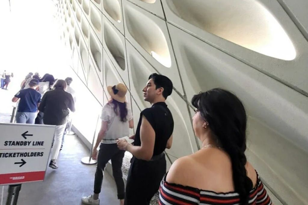 무늬가 있는 기하학적인 벽이 있는 밝은 조명의 현대적인 복도를 걷는 사람들. 한 사람은 모자를 쓰고, 다른 한 사람은 오프숄더 상의가 있습니다.