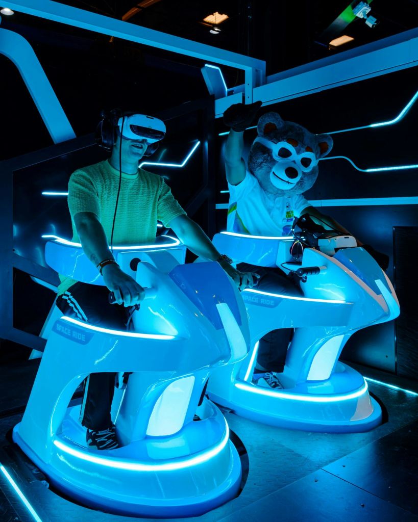 파란색 시뮬레이션 라이드에 VR 헤드셋을 착용한 두 사람이 곰 의상을 입고 미래 지향적인 네온 불빛을 배경으로 등장합니다.