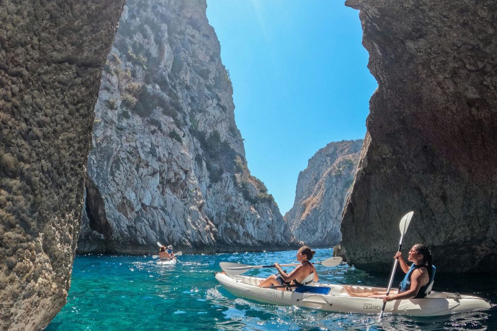 화창한 하늘 아래 가파른 바위 절벽 사이에서 맑고 푸른 바다에서 카약을 타는 사람들.