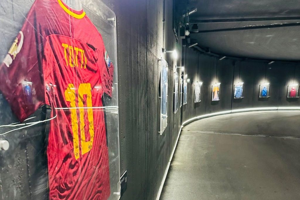 "10 Totti" 가 전면에 보이는 빨간색 저지를 포함하여 벽에 액자 스포츠 저지가 있는 희미한 조명 복도.