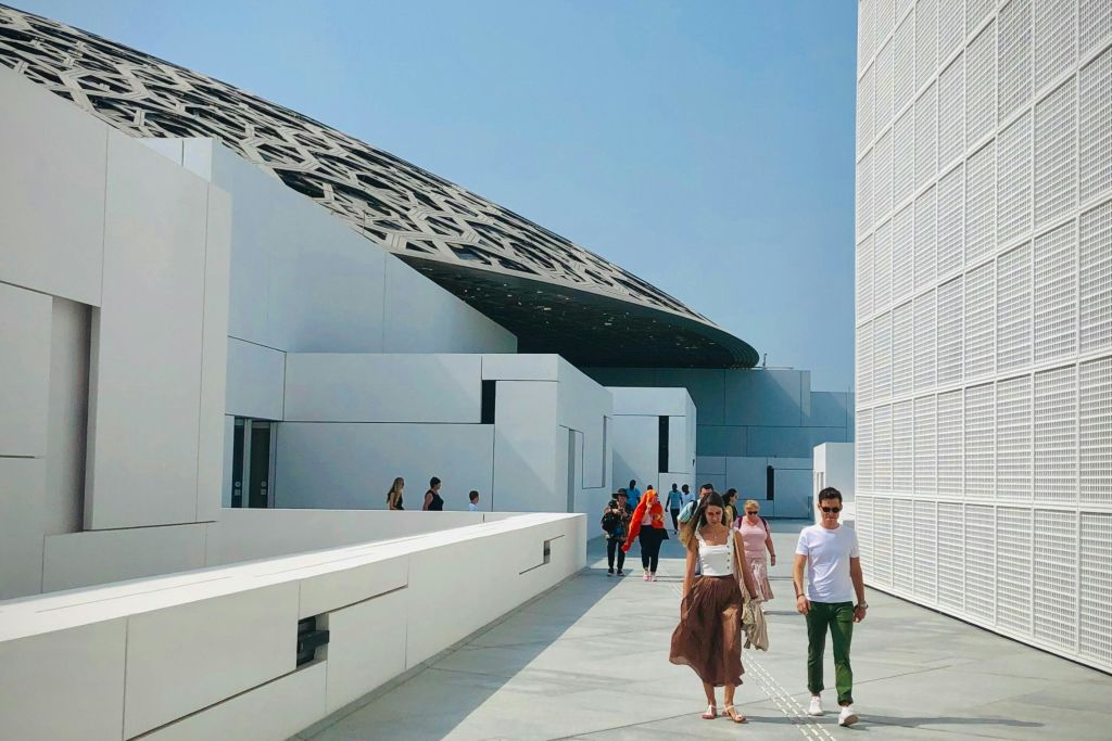 하얀 벽과 배경에 무늬가 있는 돔 구조물이 있는 현대적인 건축 통로를 걷는 사람들.