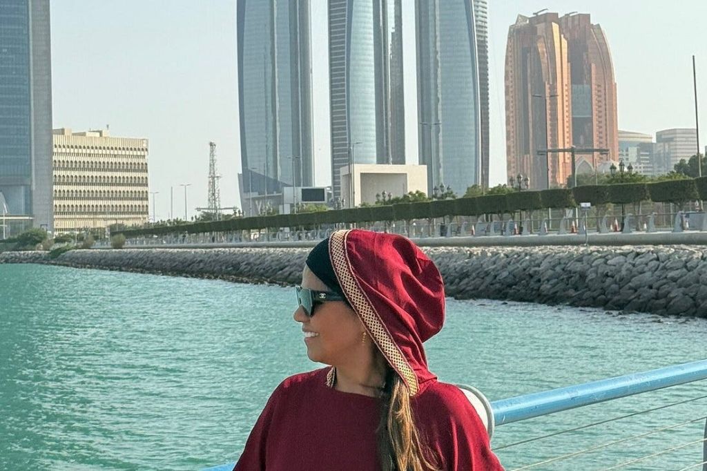 빨간색 후드가 달린 옷과 선글라스를 쓴 사람이 바닷가 근처에 서 있으며, 현대적인 고층 빌딩을 배경으로 하고 있습니다.