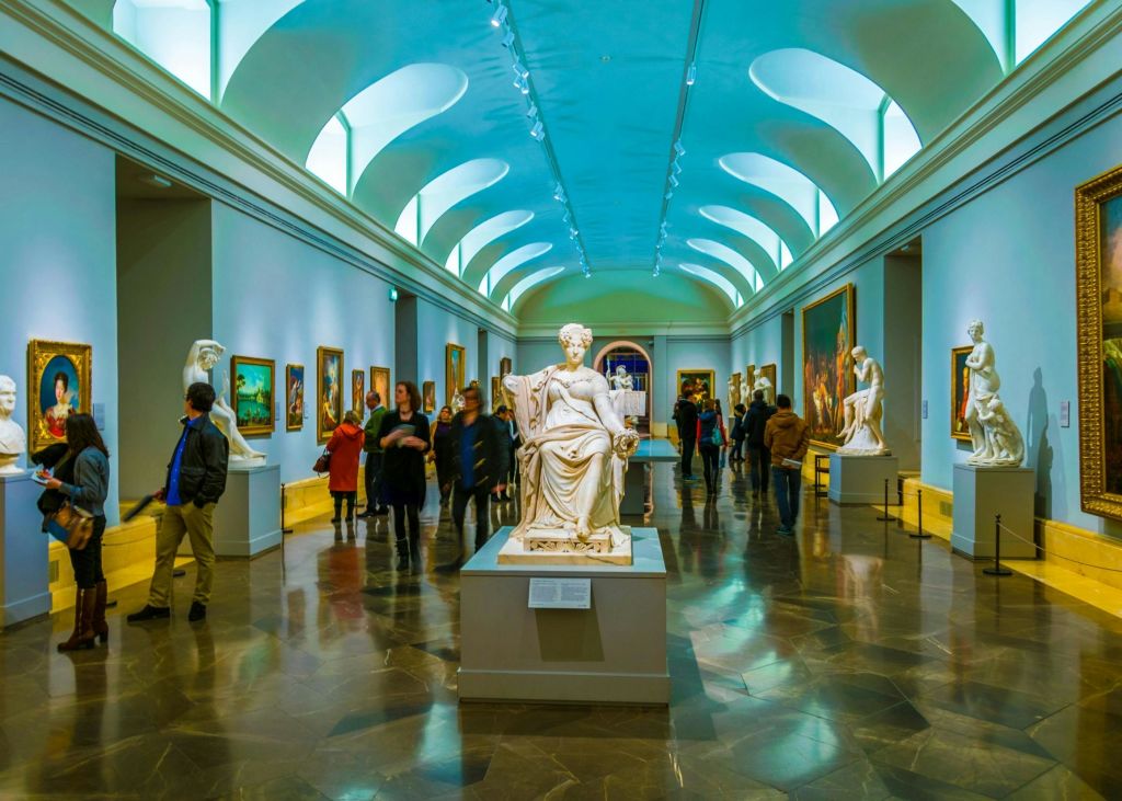 방문객들이 그림과 조각품을 감상하는 박물관 갤러리로, 아치형 천장 아래 중앙에 눈에 띄는 흰색 동상이 있습니다.