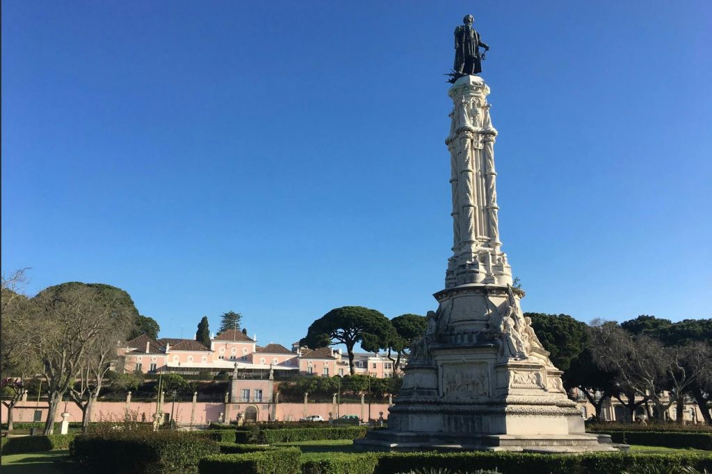 유서 깊은 분홍색 건물과 나무를 배경으로 푸른 하늘 아래 높고 화려한 조각상이 있습니다.