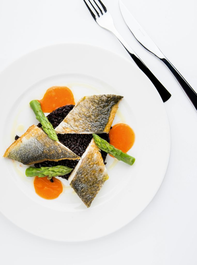 생선살, 흑미, 아스파라거스, 오렌지 소스를 식기와 함께 흰색 접시에 우아하게 플레이팅한 고급 요리입니다.