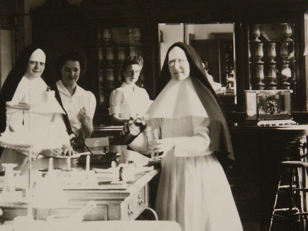 습관을 가진 수녀가 주방의 작업대에 서서 식기를 들고 있으며, 흰 유니폼을 입은 두 명의 여성이 미소를 짓고 있습니다.