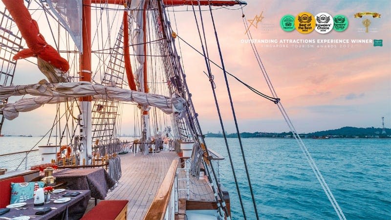 석양에 잔잔한 바다에서 즐기는 요트. 나무 데크, 돛대, 굴착기, 먼 해안선이 특징입니다. Text: "뛰어난 관광명소, 싱가포르 관광"