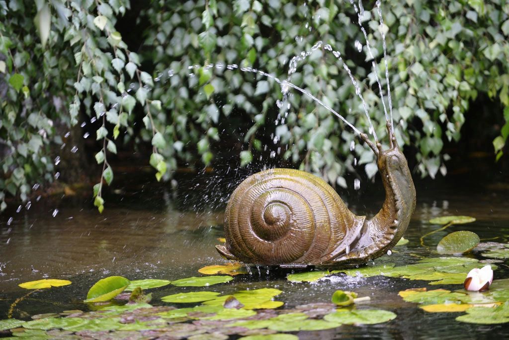 백합 패드와 매달린 단풍이 있는 연못에 있는 청동 달팽이 분수는 안테나에서 물을 뿌립니다.