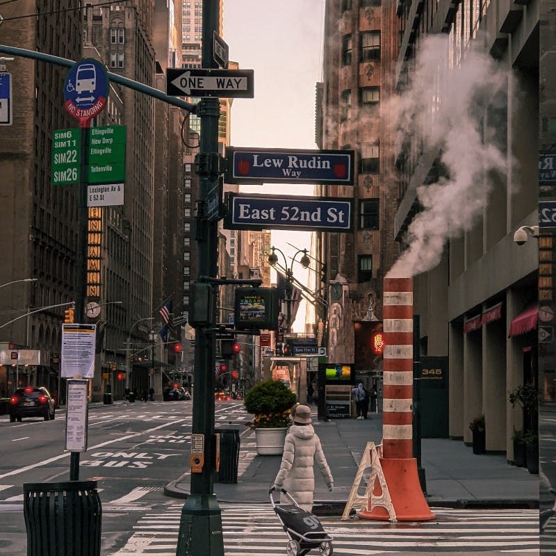 증기 배출구, "Lew Rudin Way" 와 "East 52nd St" 의 거리 표지판, 유모차를 밀고 있는 사람이 있는 뉴욕시 거리 교차로.