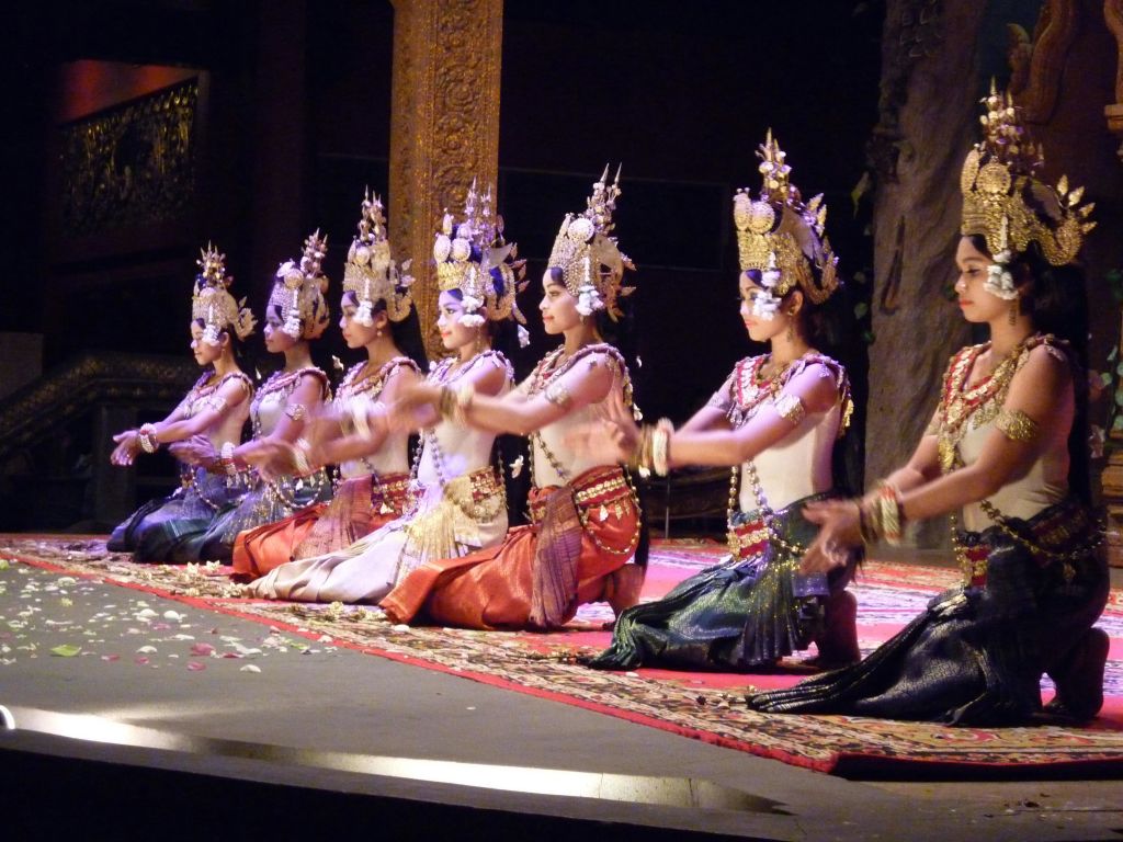 무대에서 한 줄로 앉아 여러 명의 여성이 전통적인 화려한 머리 장식과 의상을 입고 동기화된 춤을 추고 있습니다.