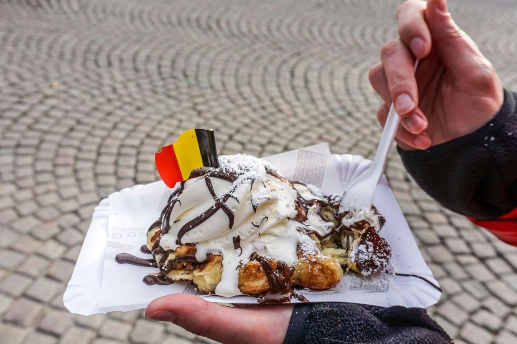 아이스크림, 초콜릿 소스, 설탕 가루, 작은 벨기에 국기를 얹은 와플이 담긴 종이 접시를 손으로 들고 있습니다.