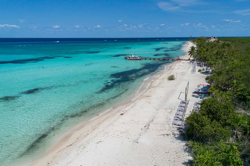 백사장이 펼쳐진 열대 해변의 공중 전망, 투명한 청록색 바다, 나무 부두, 해안가에 늘어선 빈 라운지 의자.
