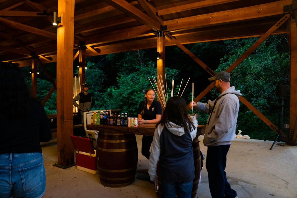통으로 만든 테이블과 음료 및 쿨러가 구비된 나무 파빌리온에 세 사람이 있습니다. 나무가 배경으로 보입니다.