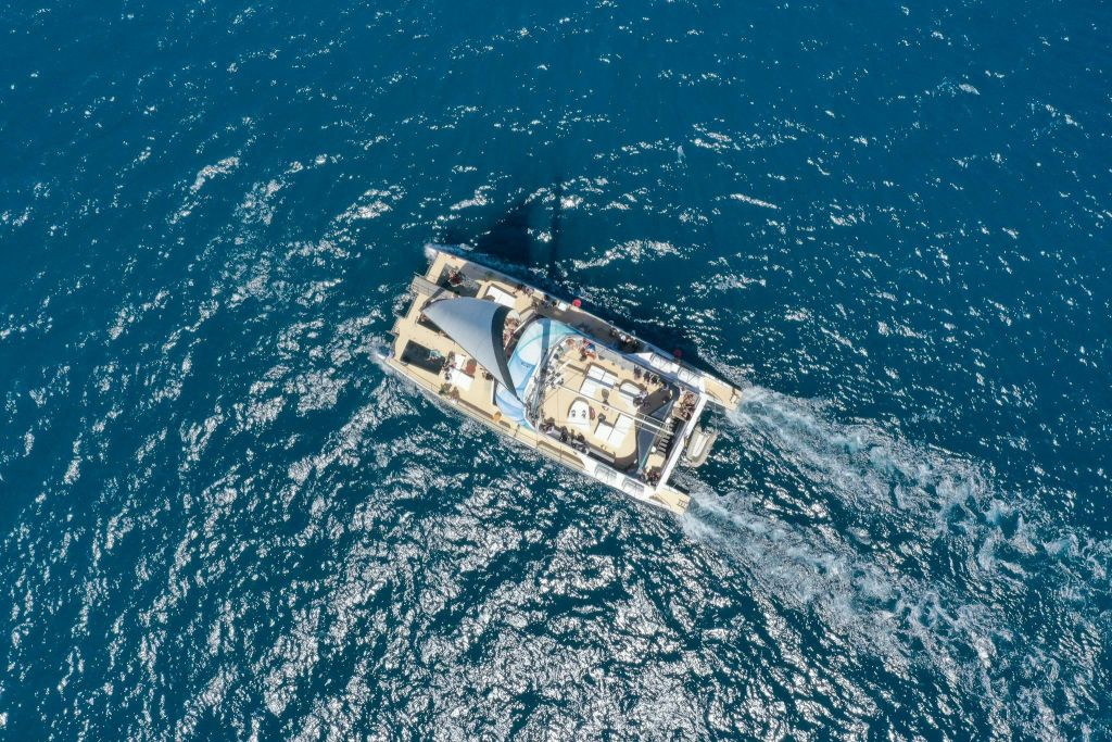 푸른 바다에서 항해하는 쌍동선의 공중 전망, 눈에 띄는 파도와 갑판 위의 사람들.