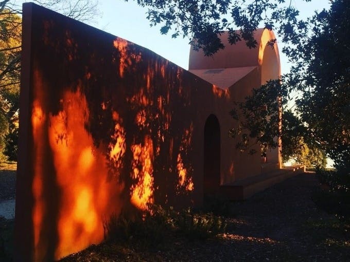 테라코타 색상의 건물로 아치형 출입구를 갖추고 있으며, 인근의 나뭇가지 사이로 햇살이 쨍쨍 내리쬐고 있습니다.