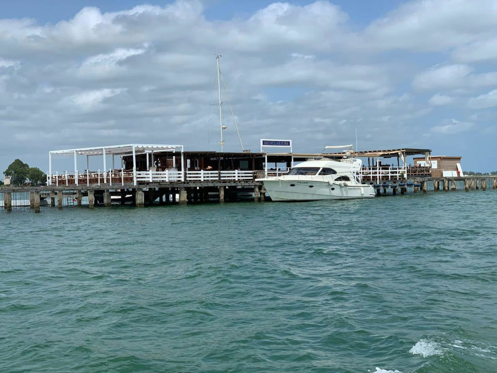 부분적으로 흐린 하늘 아래 나무 부두 근처에 흰색 요트가 정박해 있습니다. 부두에는 간판이 있는 작은 구조물이 있습니다.