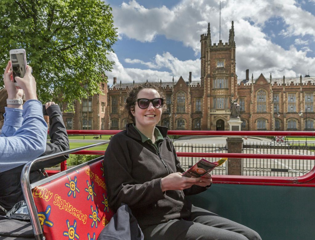 선글라스를 쓴 사람이 유서 깊은 고딕 양식 건물 근처의 2층 버스에서 안내 책자를 들고 미소를 짓고 있습니다.