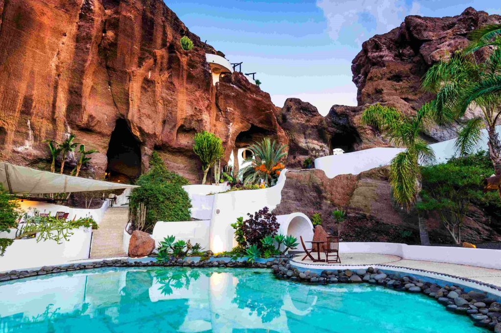 부분적으로 흐린 하늘 아래 바위 언덕에 새겨진 울창한 식물과 하얀 건물이 반사된 고요한 수영장.