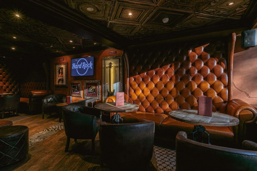 벽에 걸린 기념품 액자 위 화면에 가죽 좌석, 원형 테이블, 하드록 로고가 있는 아늑한 라운지입니다.