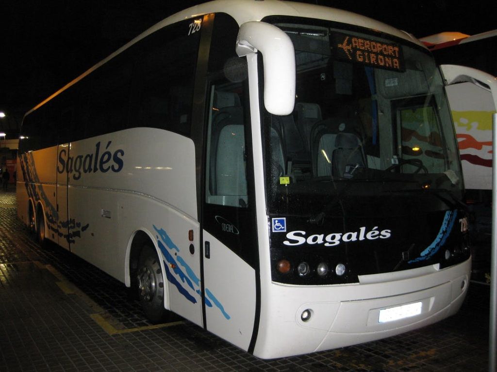전면 표지판에 "AEROPORT GIRONA" 가 표시된 화이트 사갈레스 버스는 밤에 타일 표면에 주차되어 있습니다.