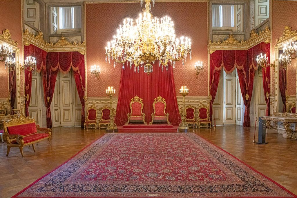 빨간색 커튼과 웅장한 샹들리에가 있는 화려한 객실로, 높이 솟은 플랫폼에 정교하게 장식된 두 개의 금색 의자가 있습니다.