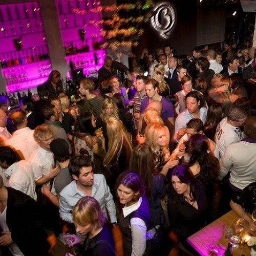 사람들이 어울리고 술을 마시는 붐비는 바입니다. 병이 있는 선반에는 분홍색 조명이 배경으로 켜져 있습니다.