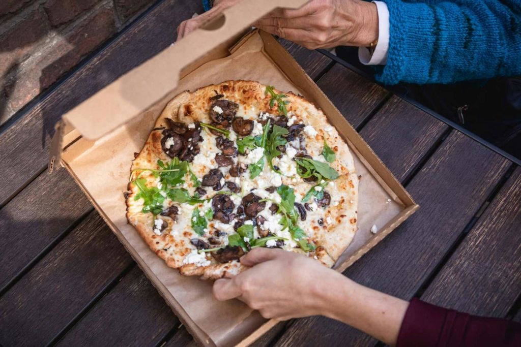 두 손이 버섯, 아루굴라, 치즈를 얹은 피자 상자를 연다. 상자는 나무 테이블 위에 놓여 있습니다.