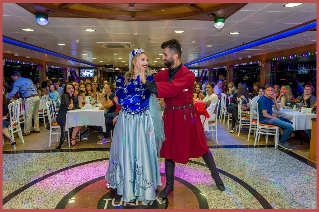 중세 의상을 입은 남녀가 보트에서 춤을 추고 있습니다. 사람들은 백그라운드에서 테이블에 앉아 그들을 지켜봅니다.