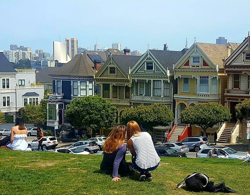 두 사람이 공원의 잔디밭에 앉아 화려한 빅토리아풍 주택을 마주하고 있습니다. 도시 건물이 배경으로 보입니다.