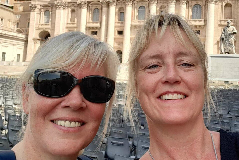 기둥과 아치가 있는 유서 깊은 건물 앞에서 미소를 짓고 있는 두 여성. 배경에는 빈 의자가 보입니다.