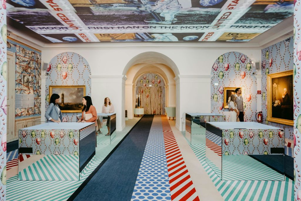 아치형 통로, 그림, 우주 비행사 천장 벽화가 있는 활기찬 무늬의 방에서 두 여성이 거울로 된 책상 위에서 대화를 나누고 있습니다.