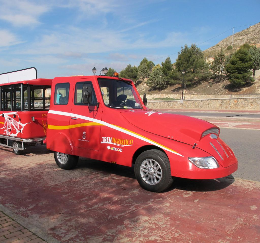 파란 하늘 아래 나무와 언덕으로 둘러싸인 포장된 지역에 마차 두 대가 주차된 빨간색 관광 열차입니다.