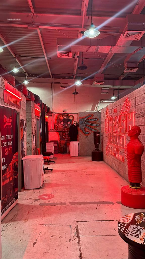 펀칭 백, 마네킹, 벽 예술품, '스매시 룸' 이라고 적힌 간판 등 빨간색 조명이 있는 방입니다.