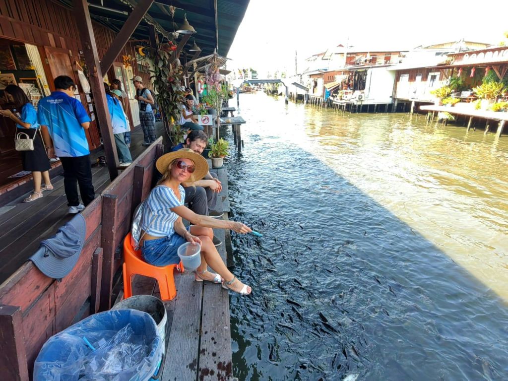모자를 쓴 여성이 물가의 오렌지색 의자에 앉아 물고기를 먹이고 있습니다. 나무 데크에 다른 여러 사람이 근처에 있습니다.