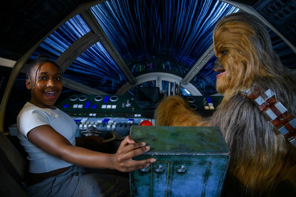 한 사람이 우주선 조종석에 앉아 미소를 짓고 있는데, 파란색의 별무늬가 있는 앞 유리창이 보입니다.