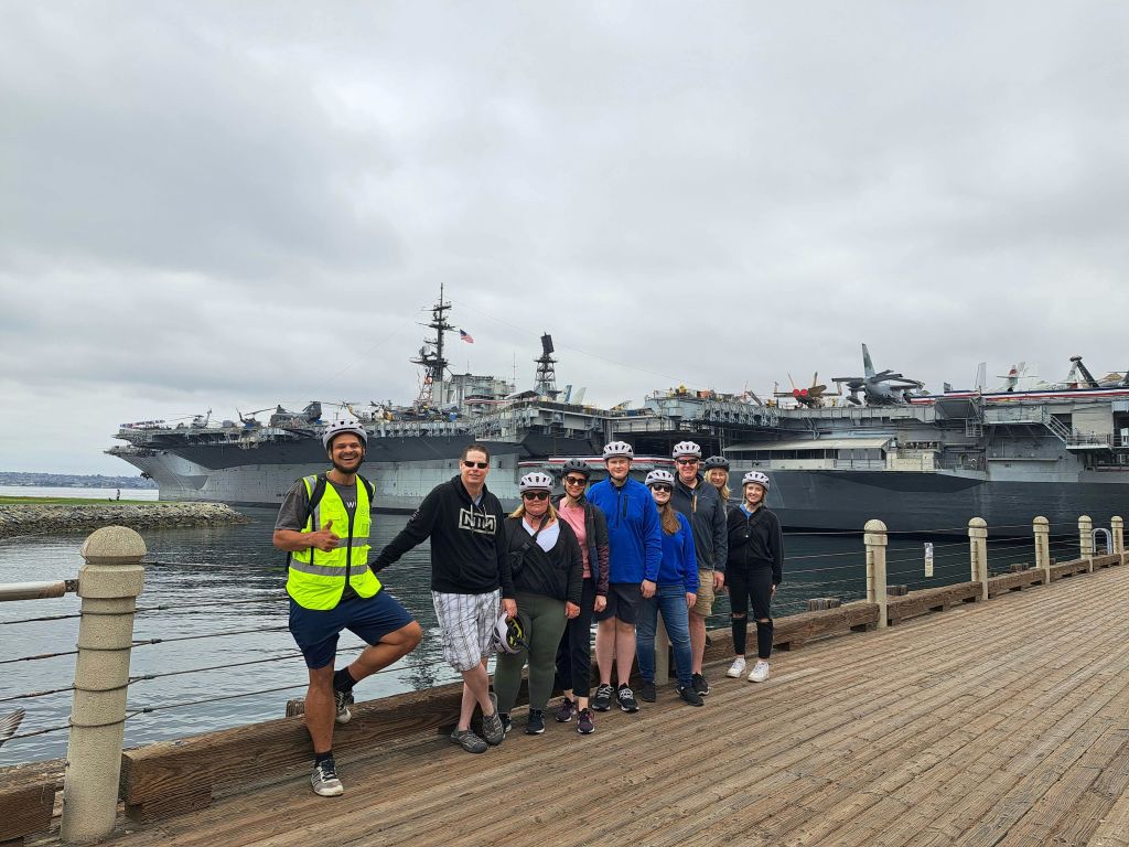 헬멧을 쓴 한 무리의 사람들이 대형 항공모함을 배경으로 보드워크에서 포즈를 취하고 있습니다.