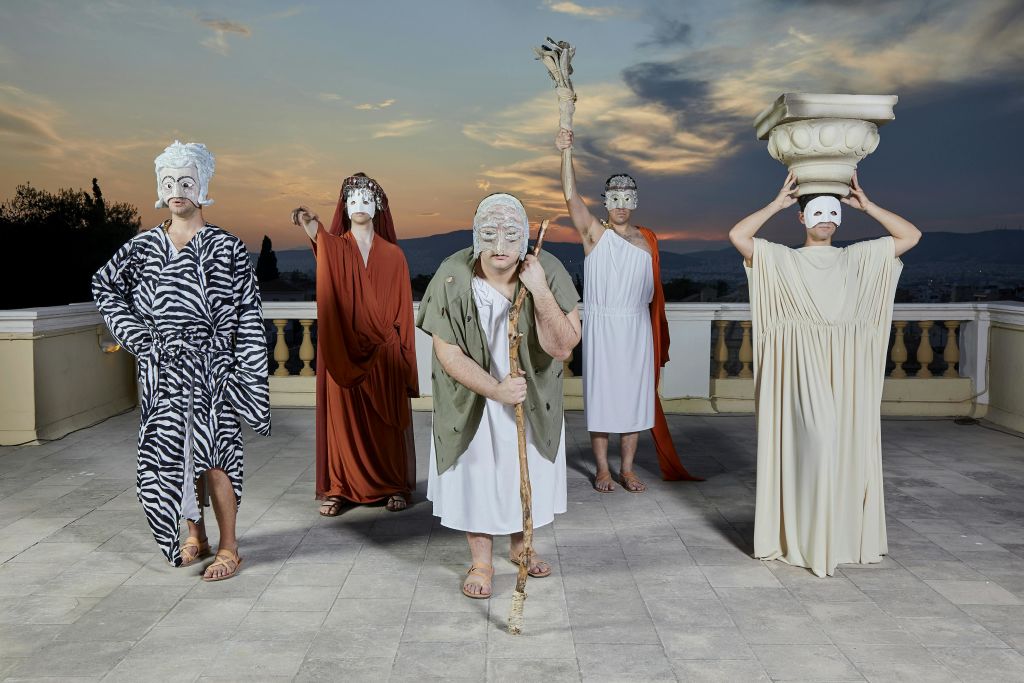 산과 극적인 석양을 배경으로 옥상에 고대 그리스 스타일의 의상과 가면을 쓴 5명이 서 있습니다.