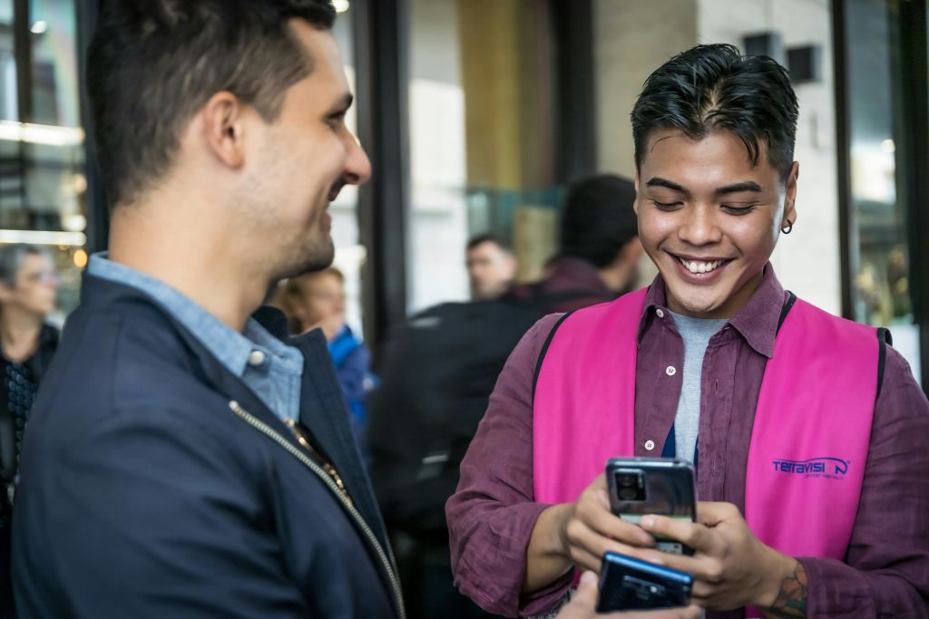 웃으며 소통하는 두 사람. 분홍색 조끼를 입은 한 사람이 휴대폰을 들고 있고, 다른 한 사람은 휴대폰을 보고 있습니다.