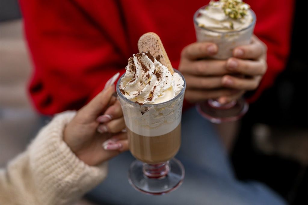 휘핑 크림, 비스코티, 장식용 토핑이 토핑된 라떼 잔을 들고 있는 두 사람. 한 사람이 빨간 스웨터를 입습니다.