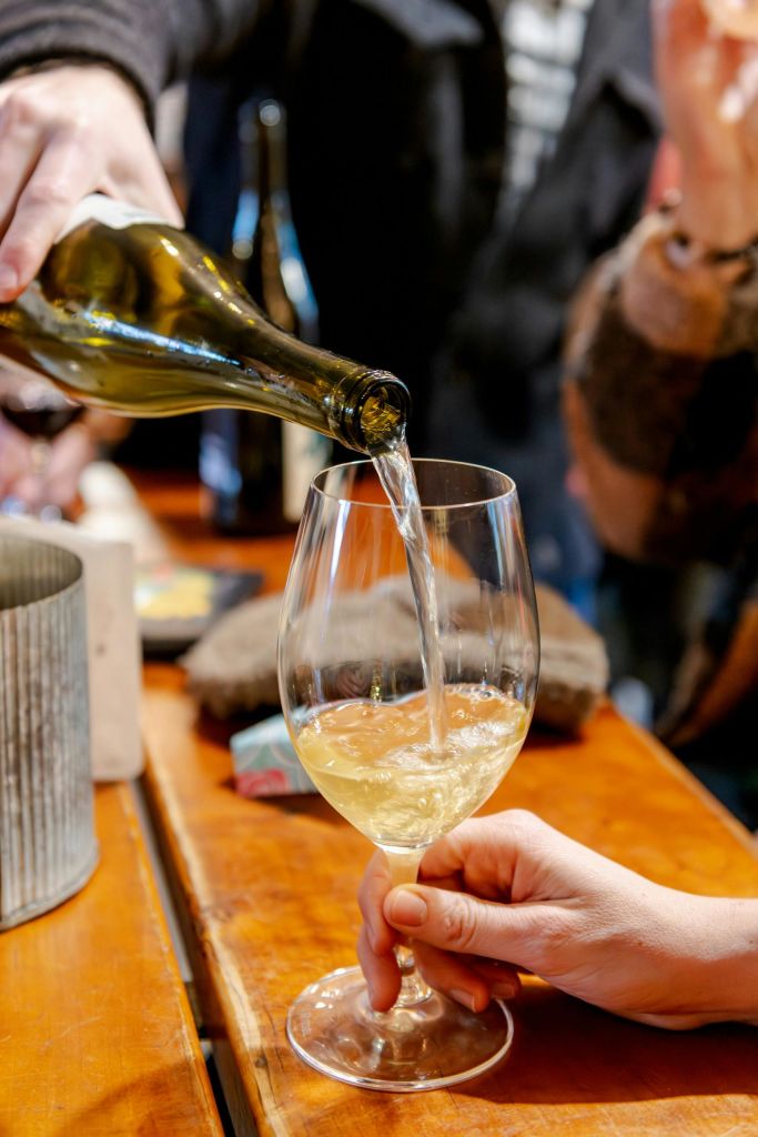 한 사람이 병에 담긴 화이트 와인을 다른 사람이 들고 있는 잔에 담아 모임에서 나무 테이블 위에 붓습니다.